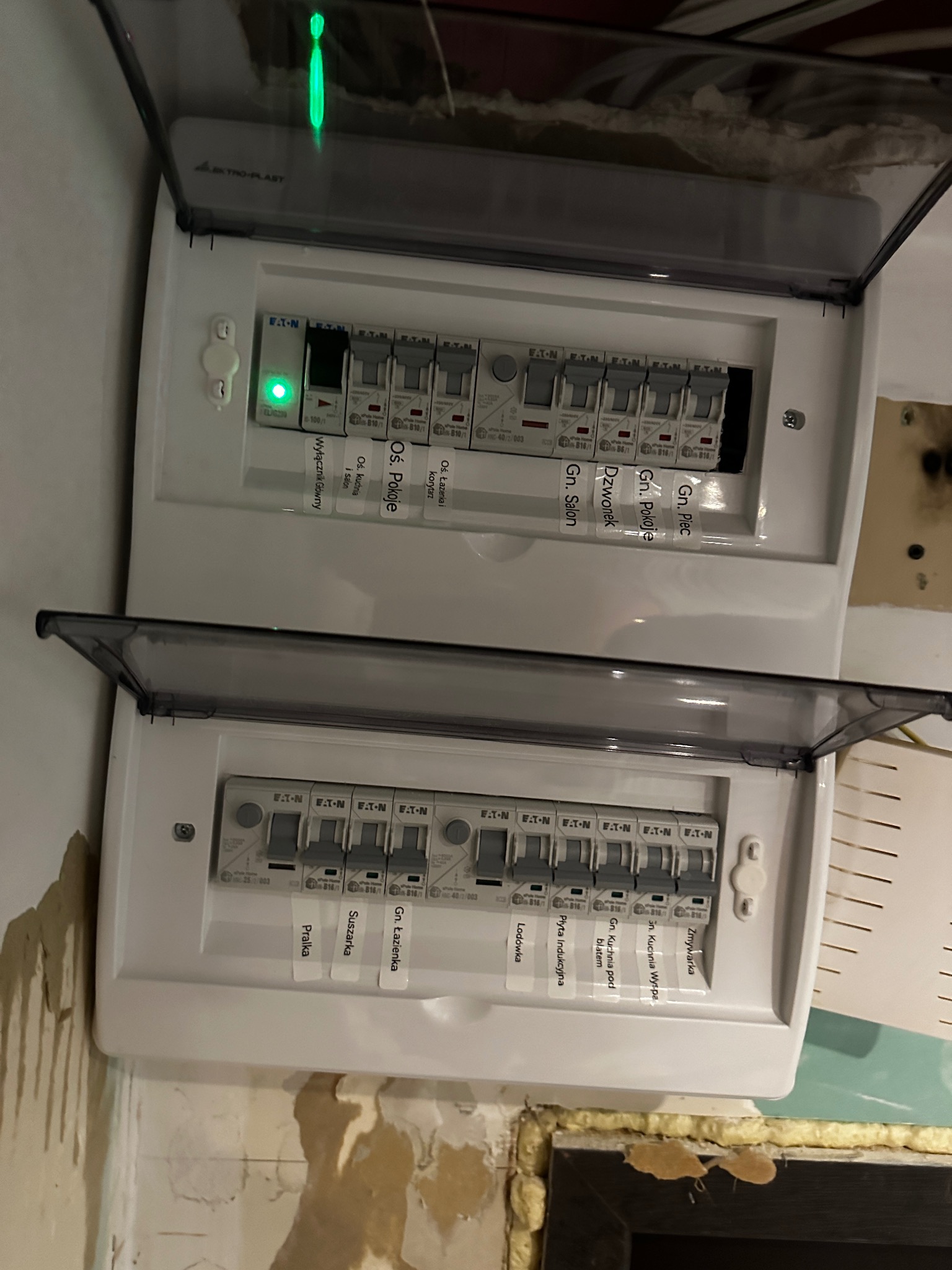Dwa białe rozdzielnice elektryczne marki Eaton z przezroczystymi osłonami, widoczne bezpieczniki z naklejkami opisującymi obwody (np. 'Wyłącznik Główny', 'Oś. Pokoje', 'Gn. Salon', 'Lodówka'...