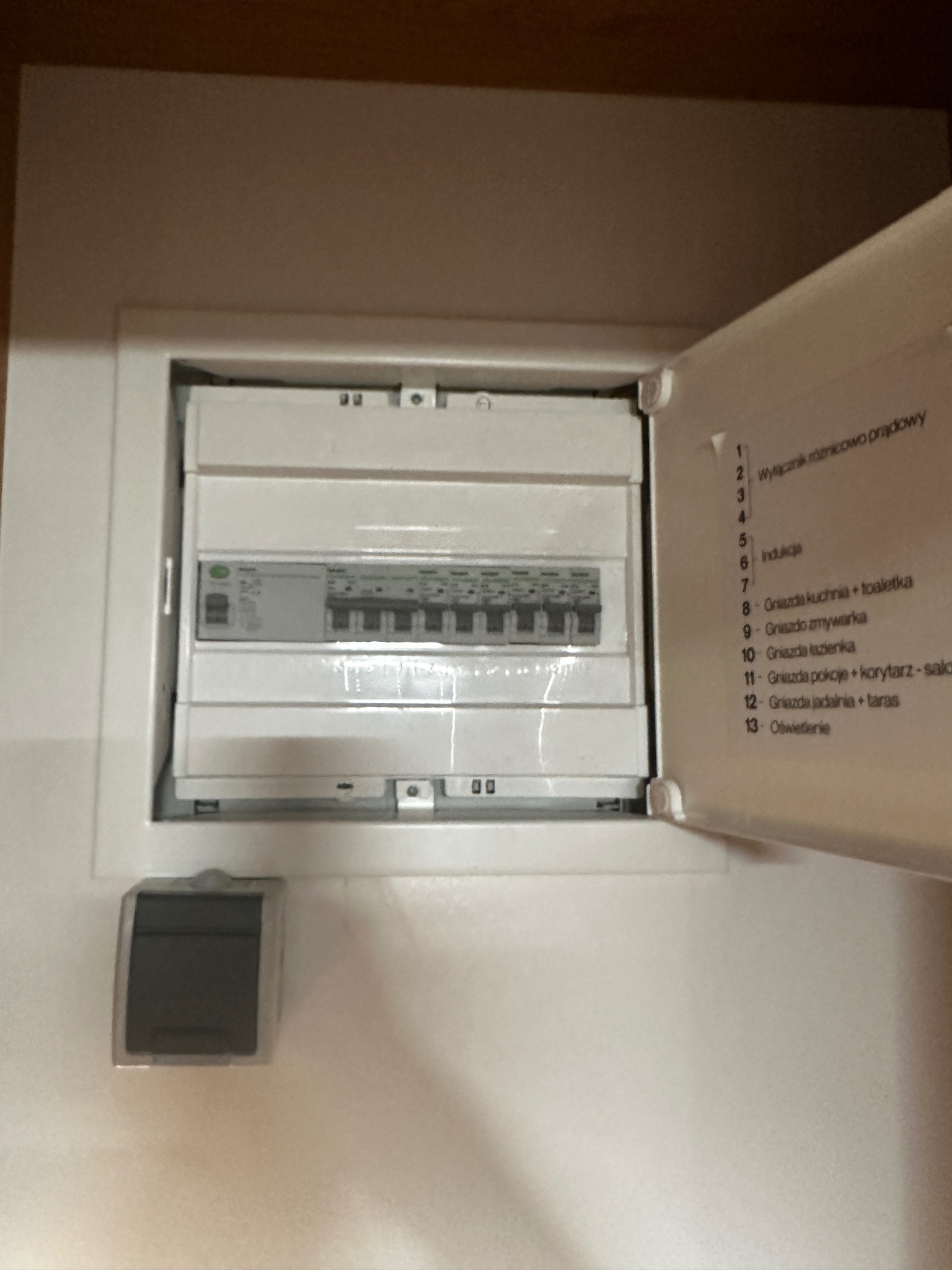 Otwarta skrzynka bezpiecznikowa z opisem obwodów, widoczne bezpieczniki i wyłączniki, obok włącznik światła.