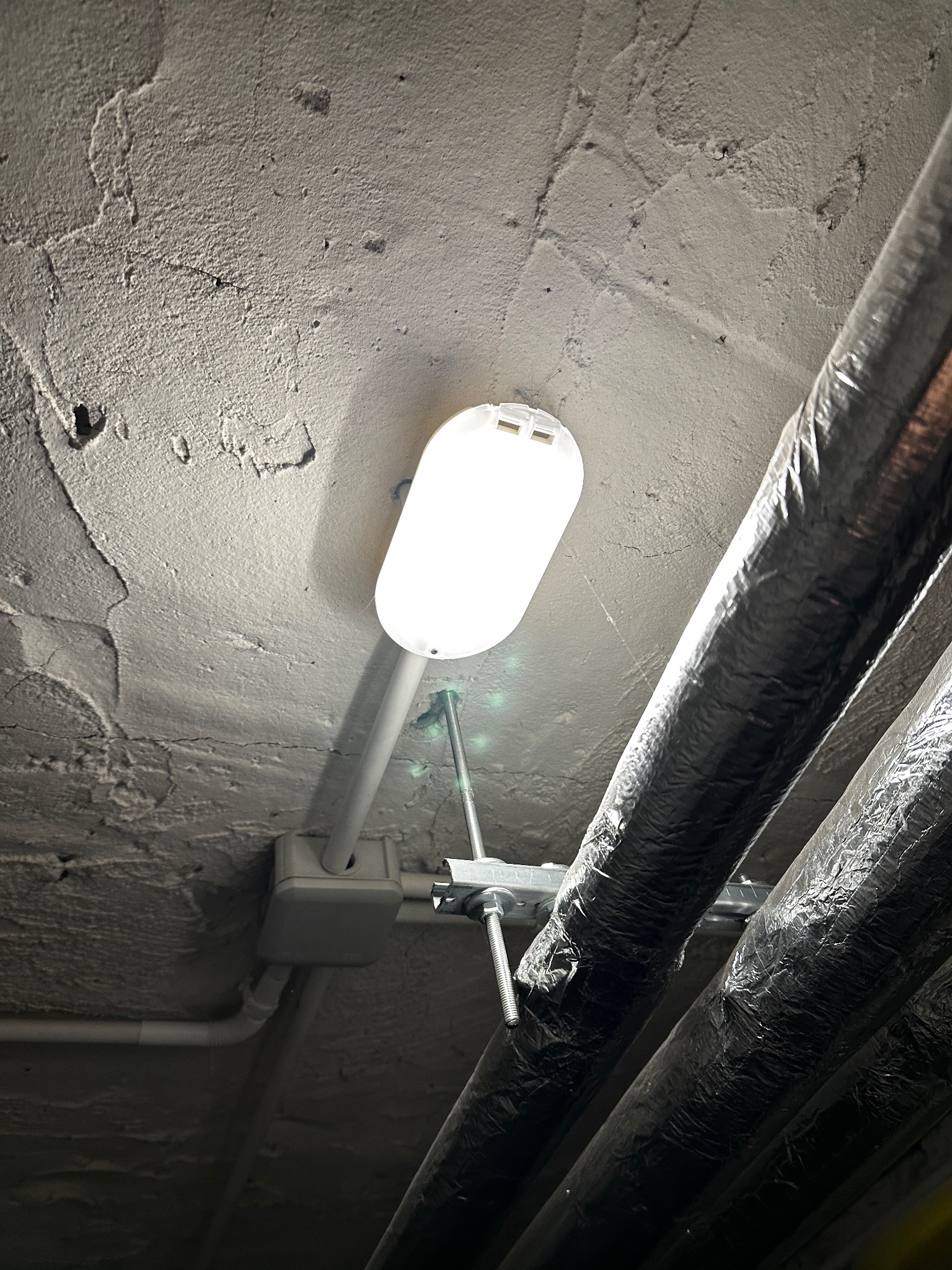 Lampa LED zamontowana na szorstkim, betonowym suficie, obok rur izolowanych folią aluminiową.