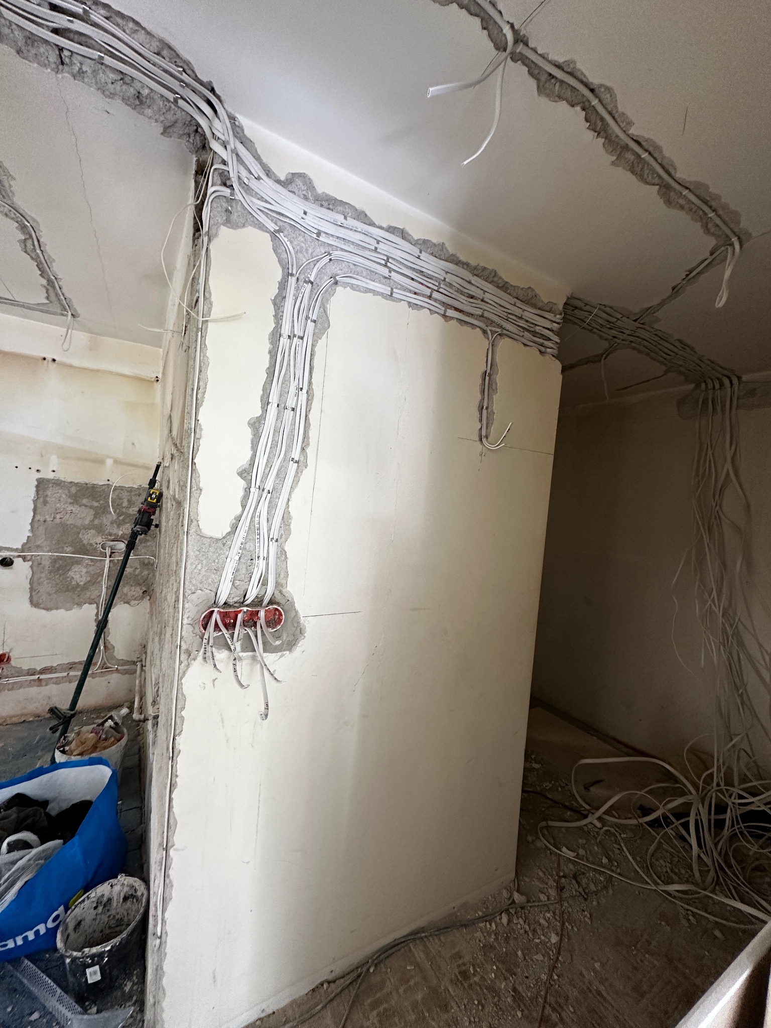 Wnętrze w trakcie remontu z bruzdami wyciętymi w ścianach i suficie pod nowe okablowanie elektryczne, widoczne przewody zabezpieczone klamrami, na podłodze gruz i narzędzia.