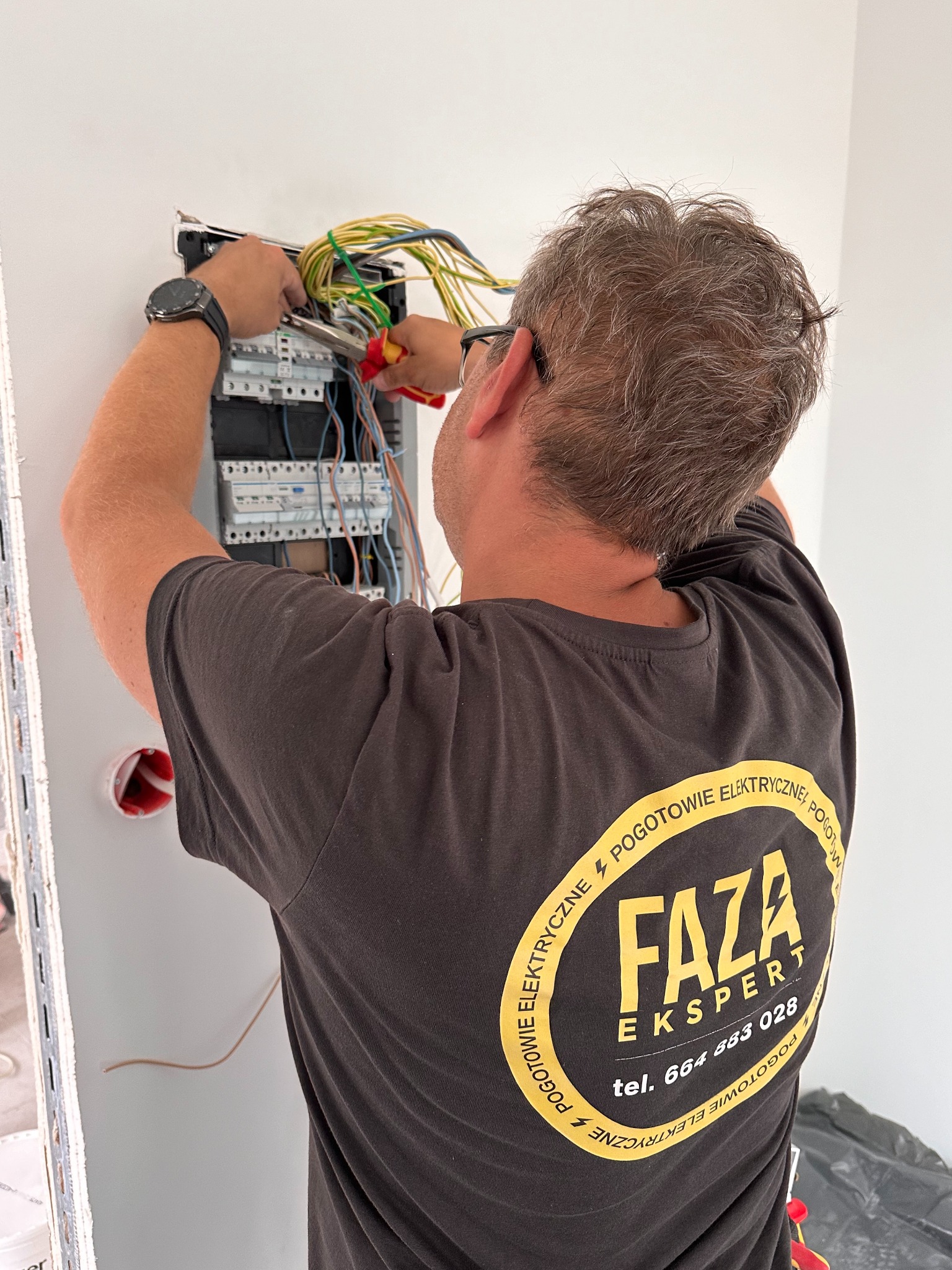 Elektryk w czarnej koszulce z żółtym logo firmy 'FAZA Ekspert' podłącza przewody do skrzynki rozdzielczej, używając kombinerek z czerwonymi uchwytami; widoczny zegarek na nadgarstku i fragment...