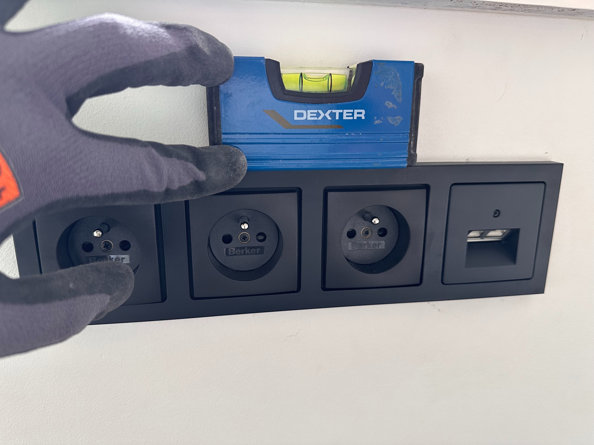 Sprawdzanie poziomu czarnej listwy z gniazdami elektrycznymi i portem USB za pomocą poziomicy Dexter przez osobę w rękawicy roboczej na tle białej ściany.