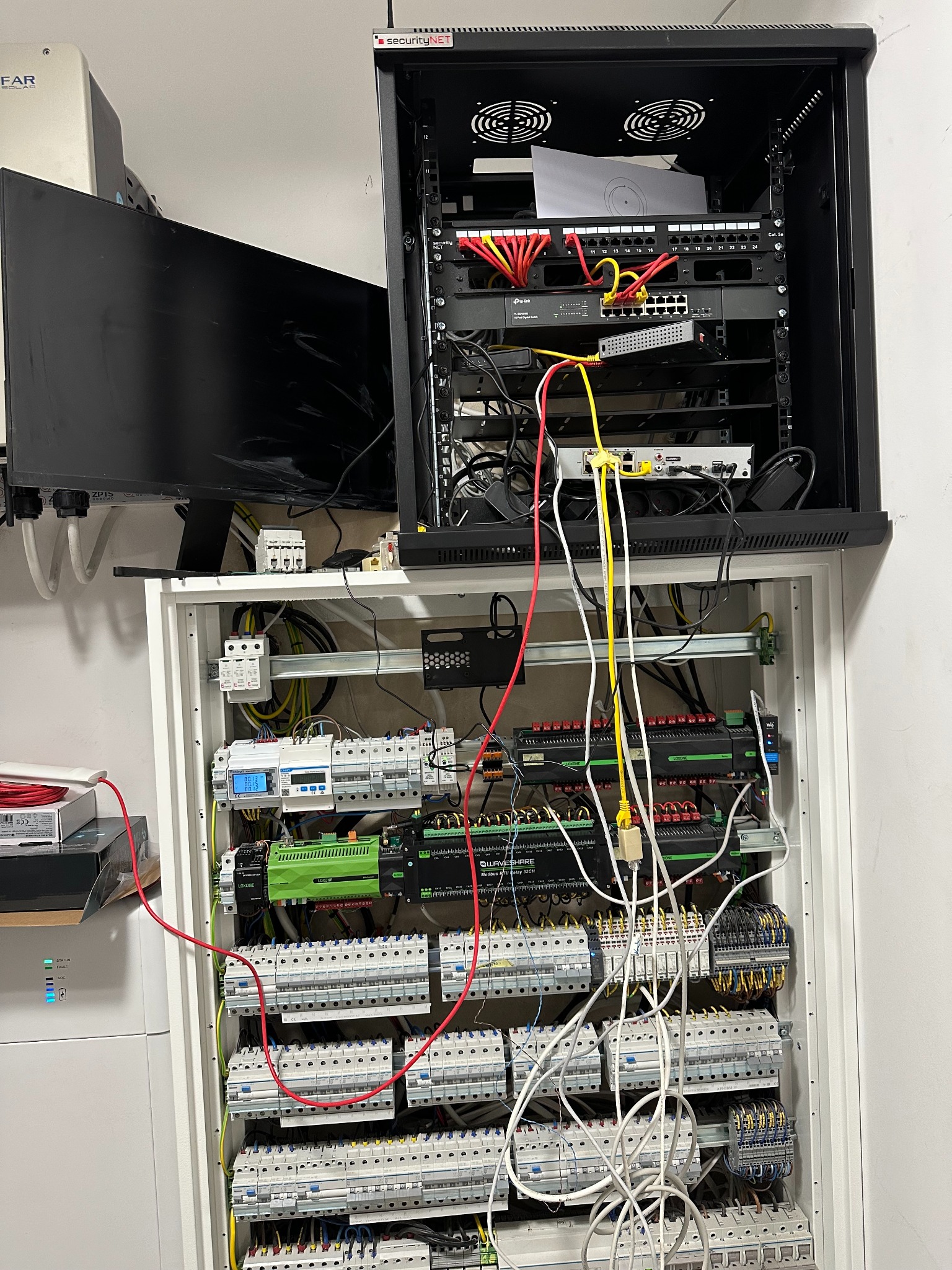 Rozdzielnia elektryczna z modułami Loxone, zabezpieczeniami, okablowaniem oraz szafa rack z routerami i przełącznikami sieciowymi, widoczne przewody Ethernet i zasilające.