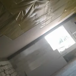 Wnętrze w trakcie prac, widoczne ocieplenie poddasza folią, zamontowane płyty gipsowo-kartonowe, surowa ściana z bloczków i okno w tle.