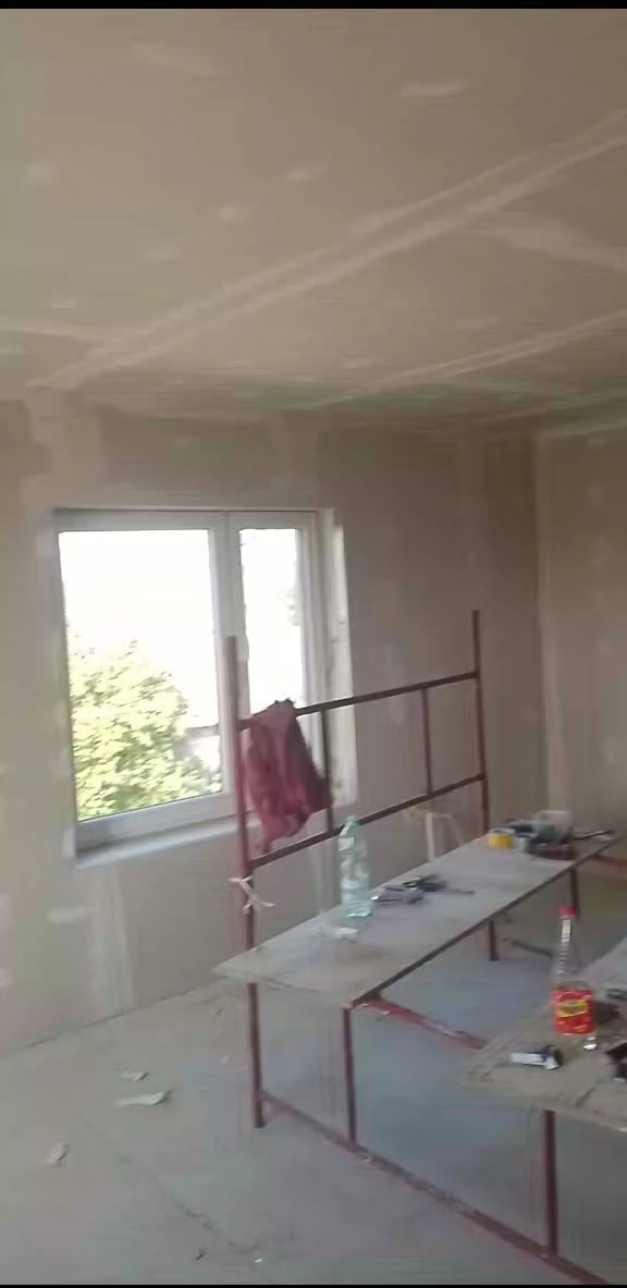 Wnętrze pokoju w trakcie prac wykończeniowych, ściany pokryte płytami gipsowo-kartonowymi, widoczne łączenia przed szpachlowaniem, prowizoryczny stół z narzędziami i napojami.