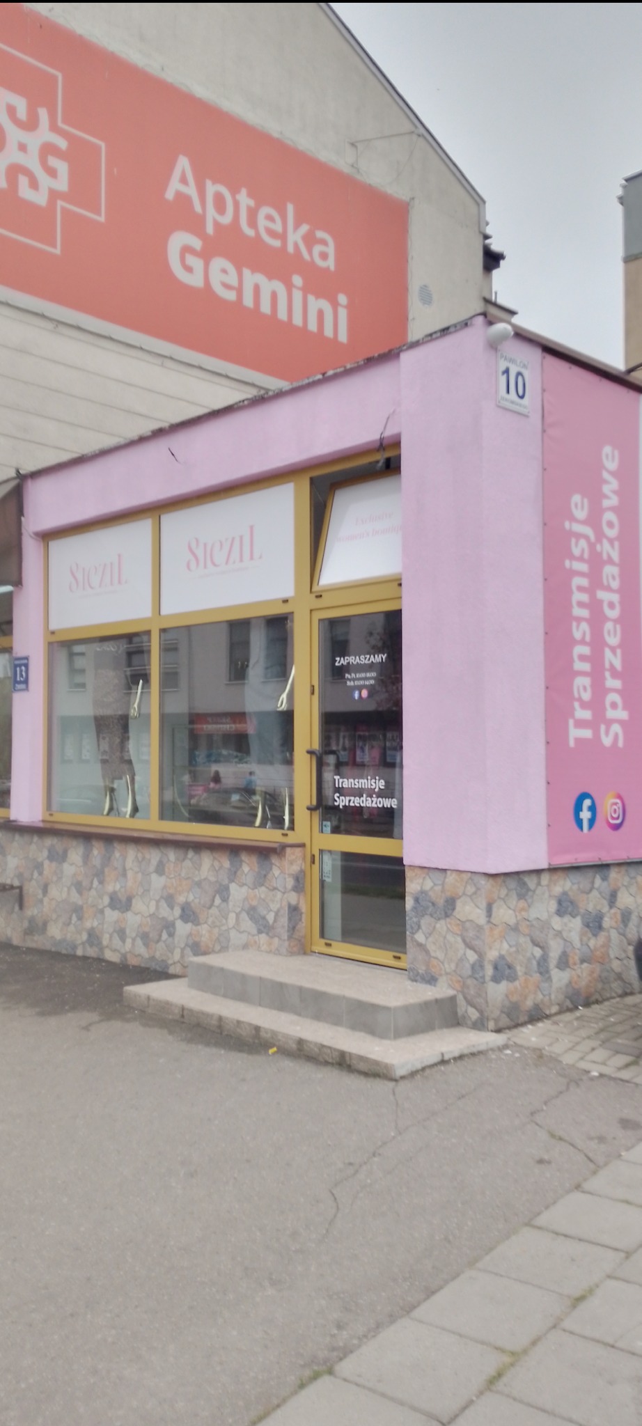 Elewacja budynku w odcieniach różu z kamienną okładziną, widoczne schody wejściowe, szyldy reklamowe i logo firmy.