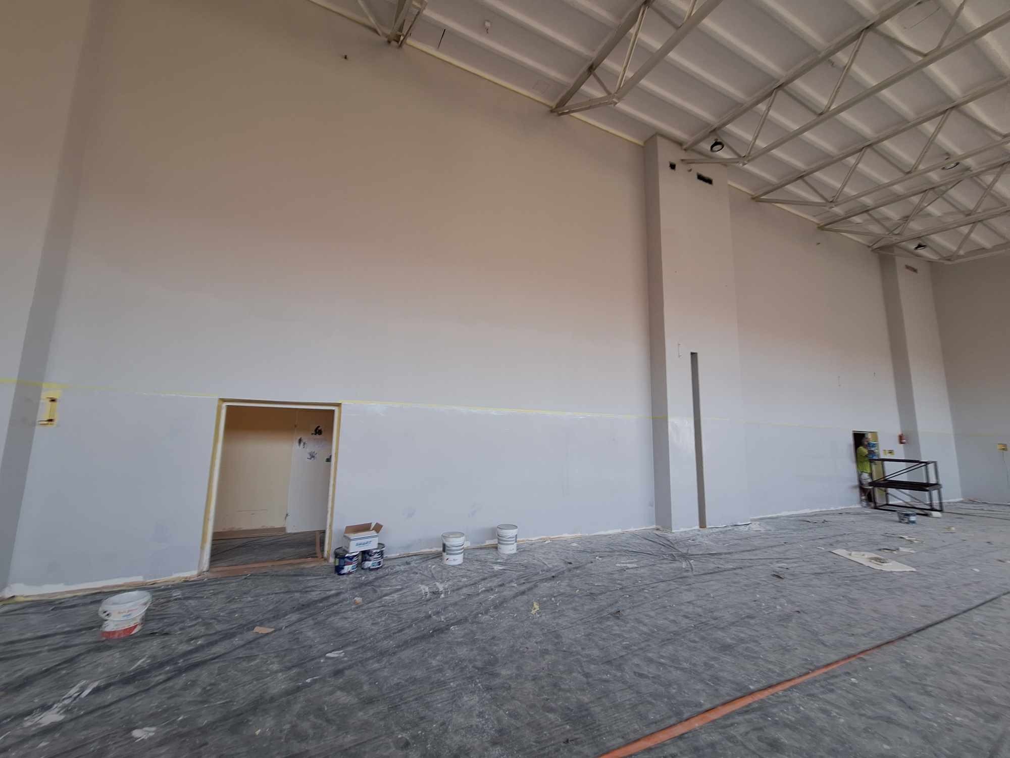 Malowanie wnętrza hali przemysłowej na szaro, widoczne puszki z farbą przy ścianie i zabezpieczona folią podłoga.