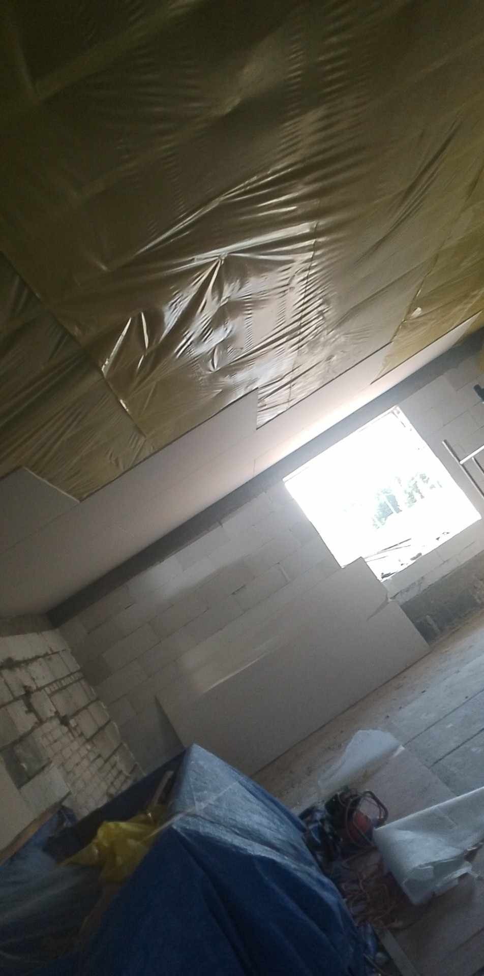 Wnętrze w trakcie prac, widoczne ocieplenie poddasza folią, zamontowane płyty gipsowo-kartonowe, surowa ściana z bloczków i okno w tle.