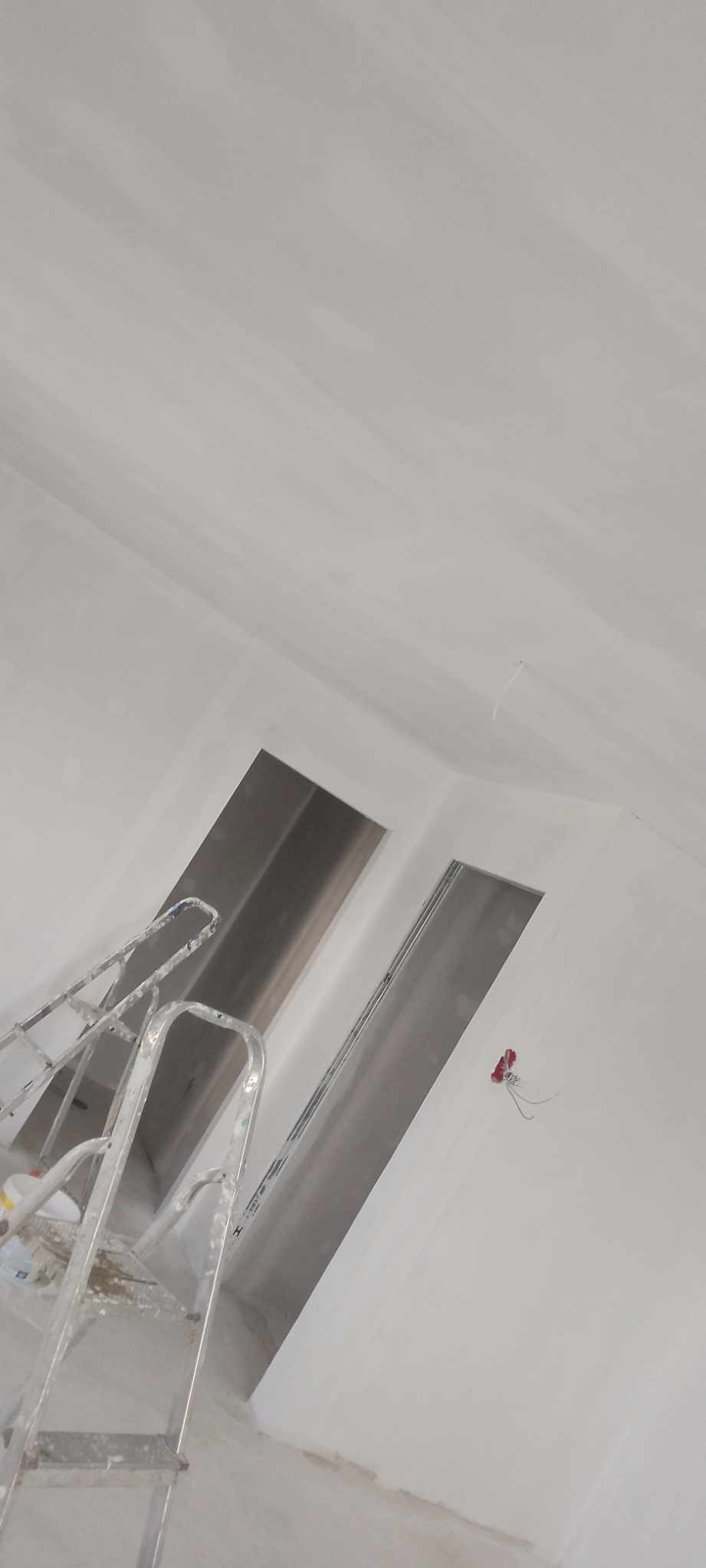 Wnętrze w trakcie prac wykończeniowych, widoczna aluminiowa drabina oparta o ścianę, sufit i ściany świeżo pomalowane na biało, widoczne kable elektryczne wystające ze ściany.