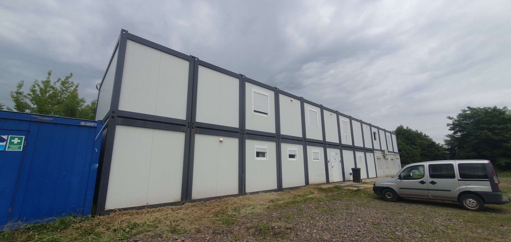 Dwu-kondygnacyjny budynek modułowy z szarymi ramami i białymi panelami, widoczne okna z roletami, obok zaparkowany srebrny van, na pierwszym planie trawa i żwir.