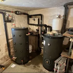 Dwa czarne akumulatory ciepła z rurami i zaworami w piwnicy z oknem dachowym i lampą sufitową, widoczne narzędzia i karton z napisem 'Folia średnia'.