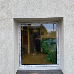 SCANDINAVIAN CONSULTING SPÓŁKA Z OGRANICZONĄ ODPOWIEDZIALNOŚCIĄ - Zamontowane okno z białymi ramami marki Drutex w elewacji z tynkiem barankiem. W szybie odbija się osoba robiąca zdjęcie oraz wnętrze pomieszczenia z zielonymi foliami ochronnymi.