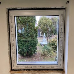 SCANDINAVIAN CONSULTING SPÓŁKA Z OGRANICZONĄ ODPOWIEDZIALNOŚCIĄ - Nowo zamontowane białe okno z widokiem na ogród z fontanną; widoczne uszczelnienie i surowy mur wokół ramy, z naklejkami producenta na ramie okiennej.