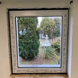 Nowo zamontowane białe okno z widokiem na ogród z fontanną; widoczne uszczelnienie i surowy mur wokół ramy, z naklejkami producenta na ramie okiennej.