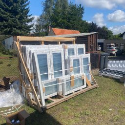 SCANDINAVIAN CONSULTING SPÓŁKA Z OGRANICZONĄ ODPOWIEDZIALNOŚCIĄ - Nowe, białe okna PCV marki Drutex, zabezpieczone folią i spięte drewnianą konstrukcją, oczekujące na montaż na trawniku przed domem, widoczne narzędzia i akcesoria montażowe w kartonie.