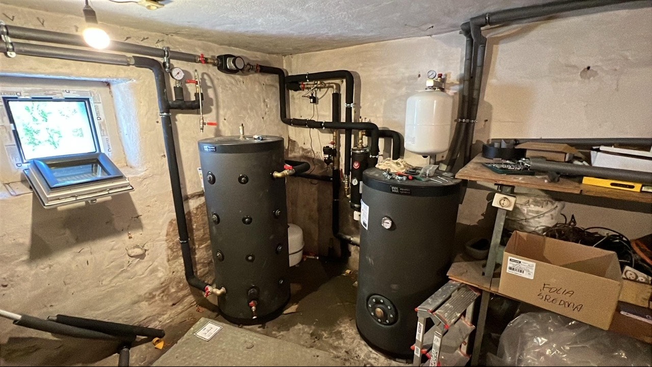 Dwa czarne akumulatory ciepła z rurami i zaworami w piwnicy z oknem dachowym i lampą sufitową, widoczne narzędzia i karton z napisem 'Folia średnia'.