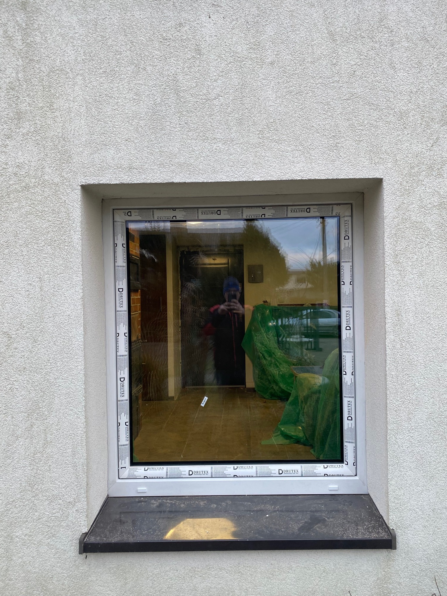 Zamontowane okno z białymi ramami marki Drutex w elewacji z tynkiem barankiem. W szybie odbija się osoba robiąca zdjęcie oraz wnętrze pomieszczenia z zielonymi foliami ochronnymi.