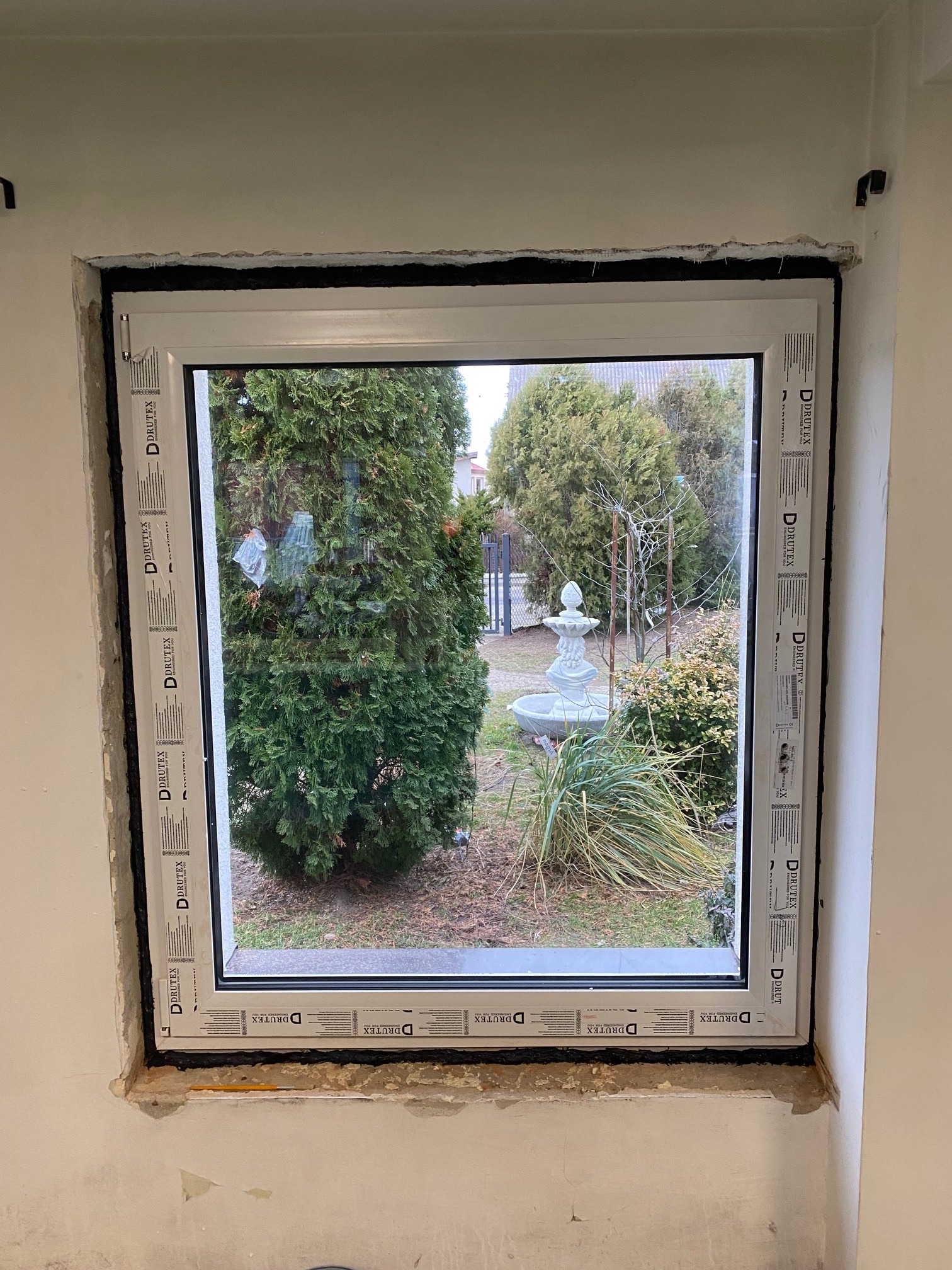 Nowo zamontowane białe okno z widokiem na ogród z fontanną; widoczne uszczelnienie i surowy mur wokół ramy, z naklejkami producenta na ramie okiennej.