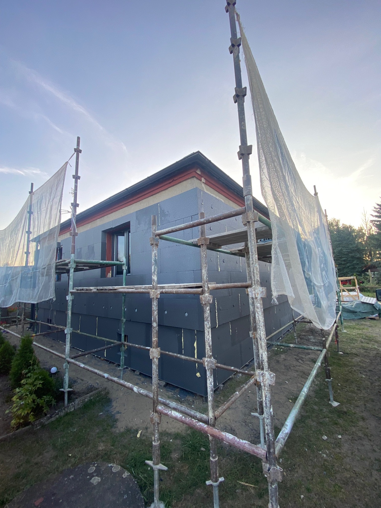 Dom w trakcie ocieplania elewacji, widoczne rusztowania, szare płyty izolacyjne z widocznymi kołkami mocującymi, okno z czarną ramą, fragment dachu z czerwoną obróbką blacharską, siatka ochronna...