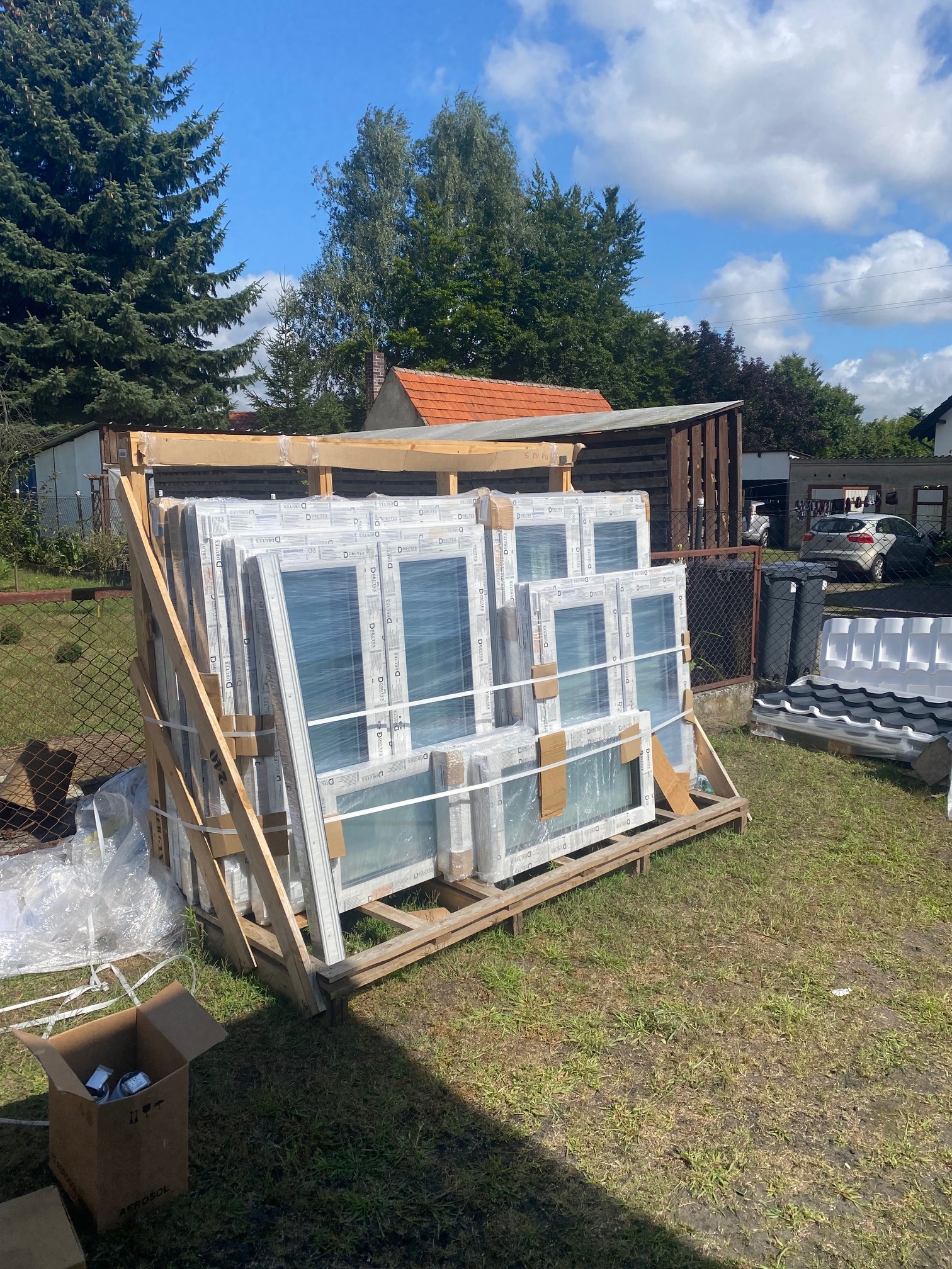 Nowe, białe okna PCV marki Drutex, zabezpieczone folią i spięte drewnianą konstrukcją, oczekujące na montaż na trawniku przed domem, widoczne narzędzia i akcesoria montażowe w kartonie.