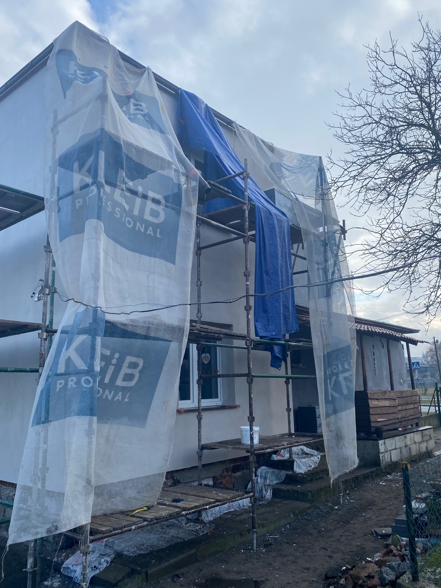 Dom w trakcie ocieplania elewacji, rusztowania częściowo zakryte banerami reklamowymi firmy, widoczny fragment ocieplonej ściany, w tle sąsiedni budynek i drzewo bez liści, na ziemi gruz.