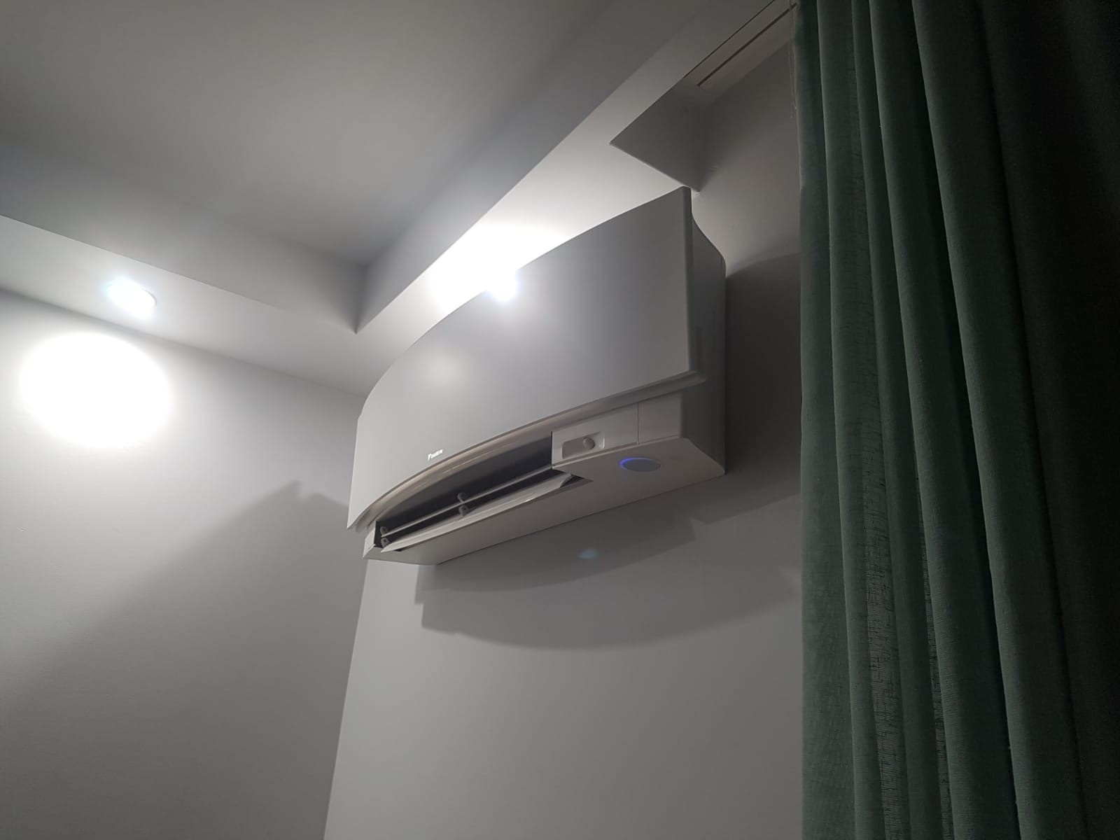 Biała klimatyzacja ścienna Daikin zamontowana w pomieszczeniu, widoczna część zielonej zasłony po prawej stronie.