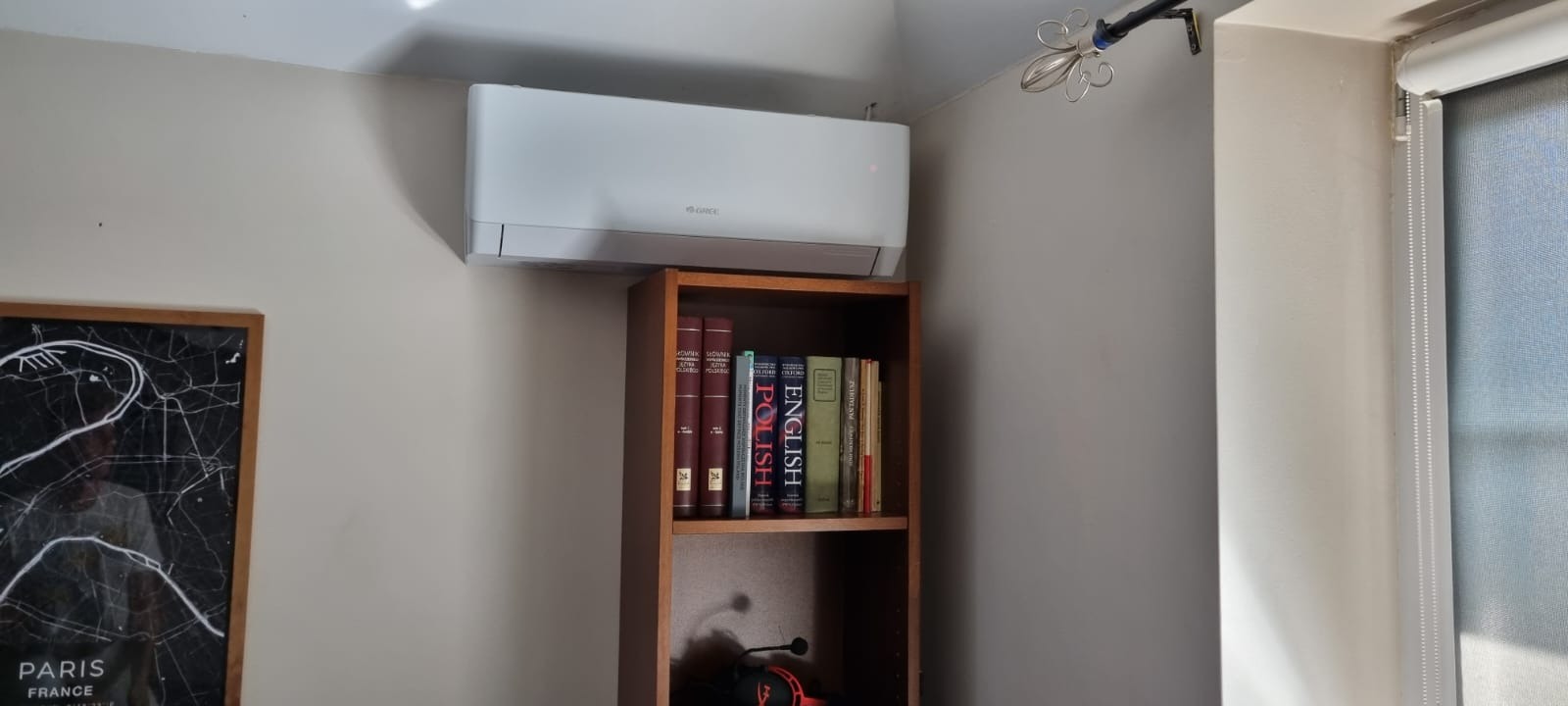 Biały klimatyzator ścienny marki Gree zamontowany nad drewnianym regałem z książkami w pokoju z szarą ścianą i zasłoniętym oknem.