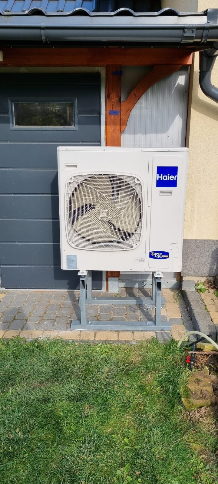 Zewnętrzna jednostka klimatyzacji Haier zamontowana na metalowym stelażu przy ścianie budynku z szarą bramą garażową i zielonym trawnikiem.