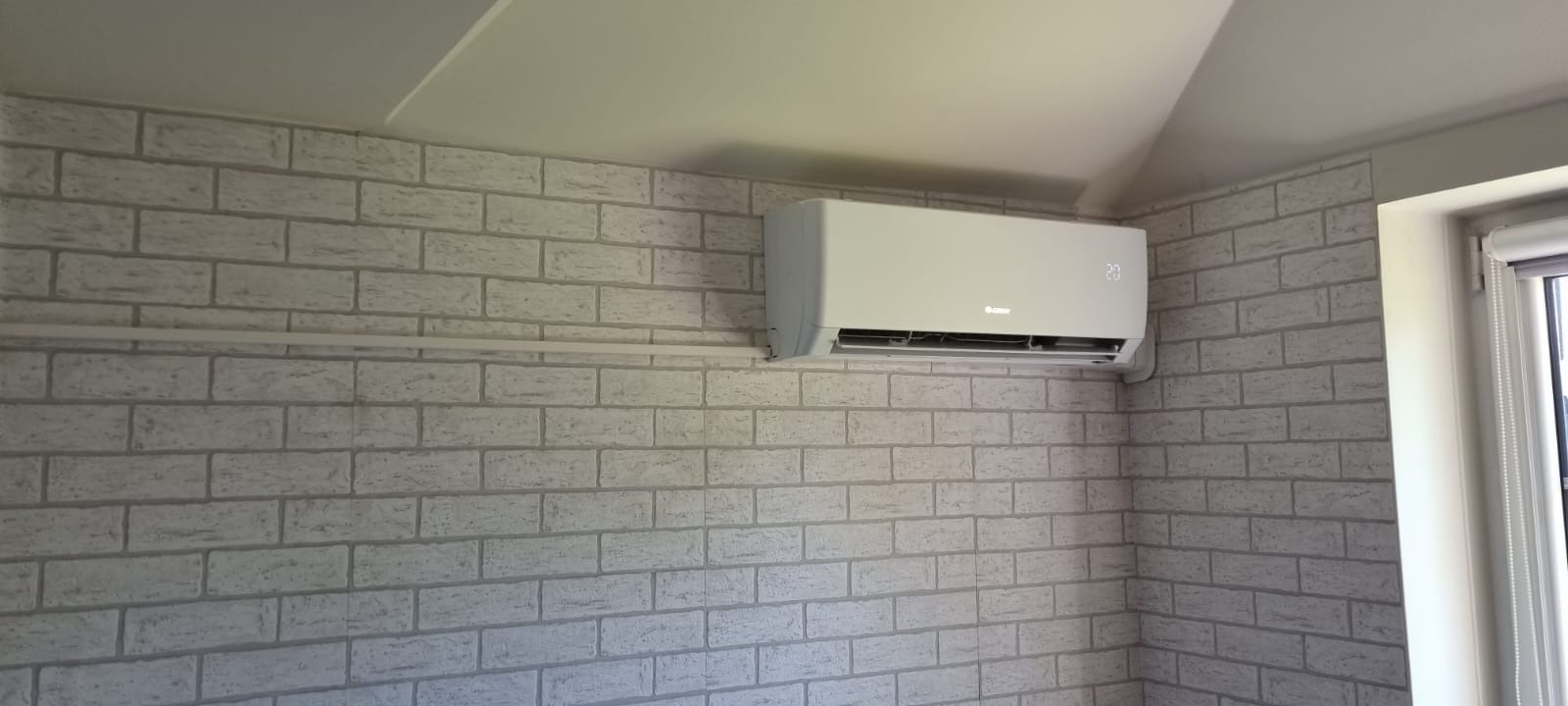 Biały klimatyzator marki Gree zamontowany na ścianie z tapetą imitującą cegłę w pokoju na poddaszu, widoczny wyświetlacz temperatury.