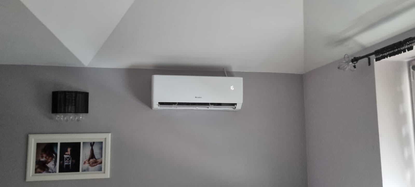 Biały klimatyzator ścienny marki Gree z wyświetlaczem temperatury, zamontowany w szarym pokoju z dekoracyjną lampą i ramką ze zdjęciami na ścianie.