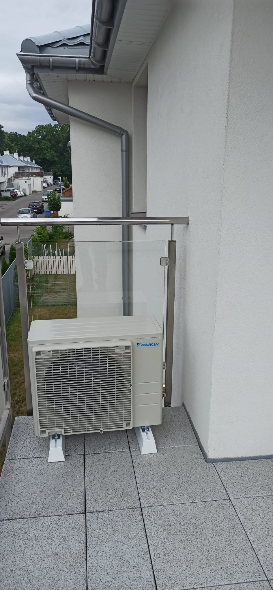 Jednostka zewnętrzna klimatyzacji Daikin zamontowana na balkonie z szarą płytką i szklaną balustradą, widok na osiedle domów.