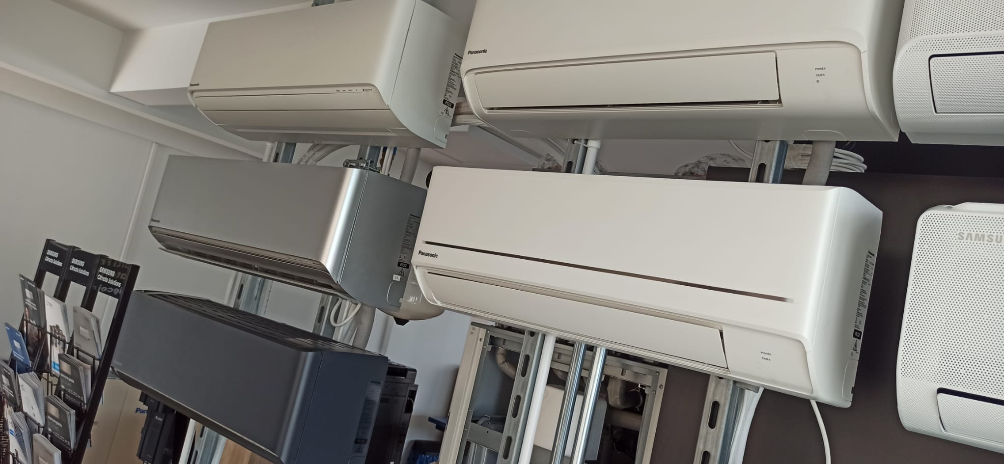 Ekspozycja różnych modeli klimatyzatorów Panasonic i Samsung w salonie, widoczne jednostki wewnętrzne w kolorach białym, szarym i granatowym, zamontowane na konstrukcji wsporczej z widocznymi...