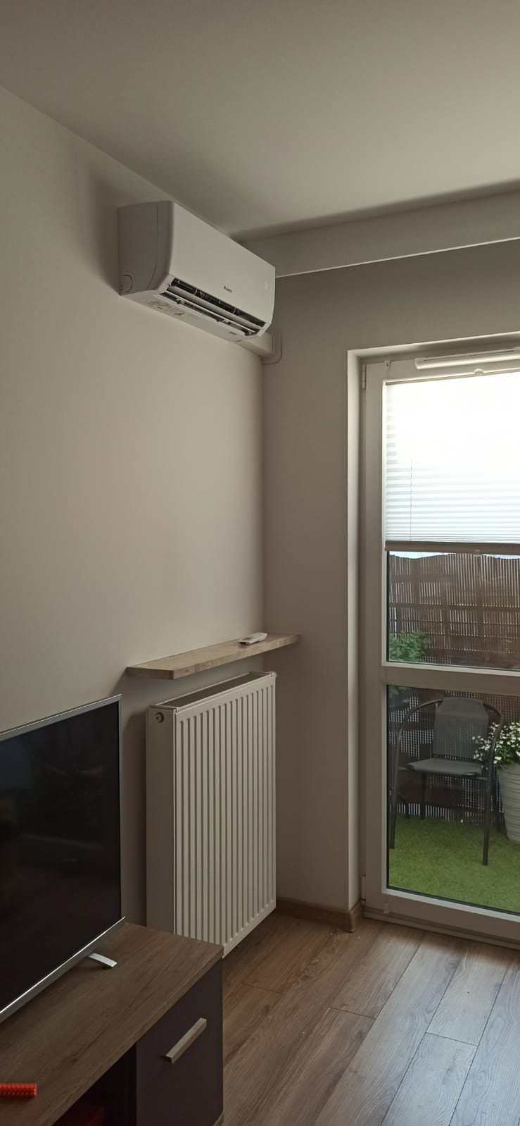 Nowoczesny klimatyzator ścienny w jasnym pokoju z grzejnikiem pod półką, widok na balkon z krzesłem i zieloną trawą.