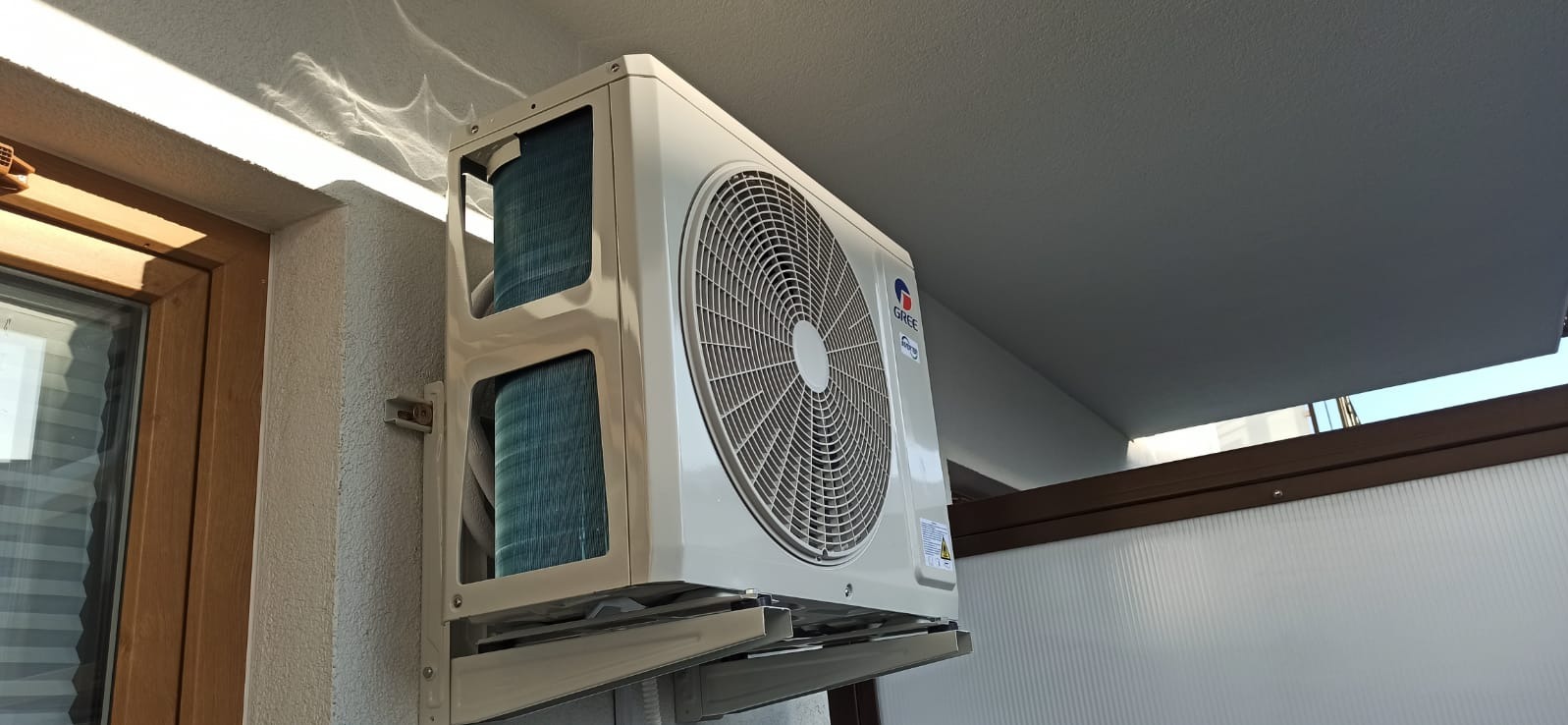 Zewnętrzna jednostka klimatyzacji marki Gree zamontowana na ścianie budynku, widok z dołu, widoczne żaluzje i logo producenta.