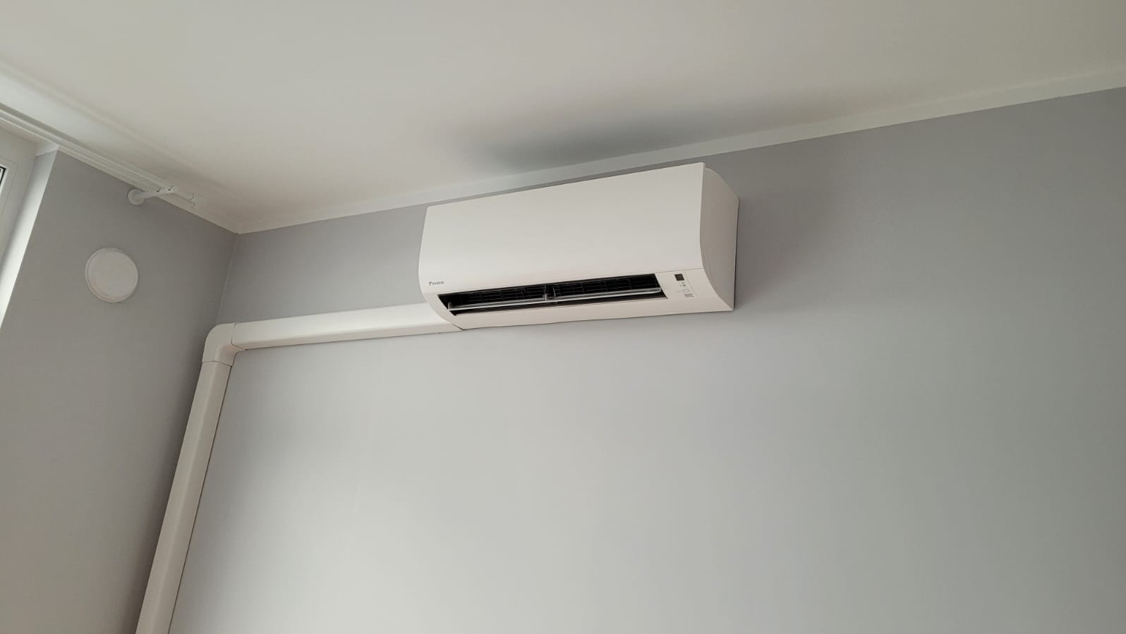 Biały klimatyzator Daikin zamontowany na szarej ścianie w pomieszczeniu z białym sufitem i widocznymi białymi rurami instalacyjnymi.