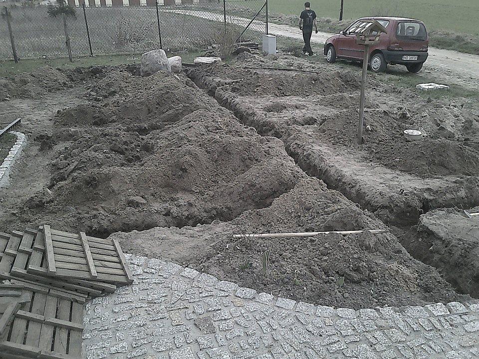 Wykonane wykopy pod system nawadniania ogrodu, widoczny fragment kostki brukowej, sterta desek, w tle osoba i czerwony samochód osobowy.