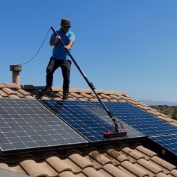 SolCare Nicola Danelczak - Osoba w okularach przeciwsłonecznych czyści panele słoneczne na dachu domu za pomocą szczotki na długim wysięgniku, na tle błękitnego nieba i zabudowań.