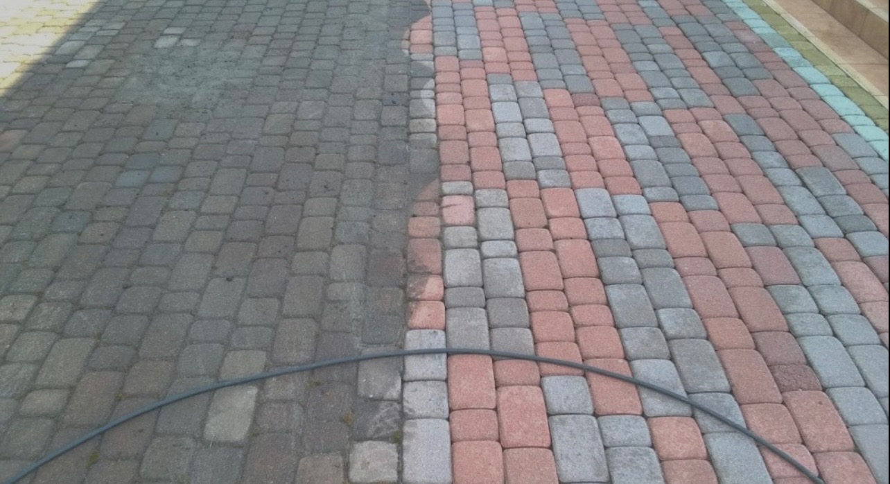 Częściowo wyczyszczona kostka brukowa w odcieniach szarości i czerwieni, widoczna różnica w kolorze i czystości, z fragmentem węża ciśnieniowego w dolnej części kadru.