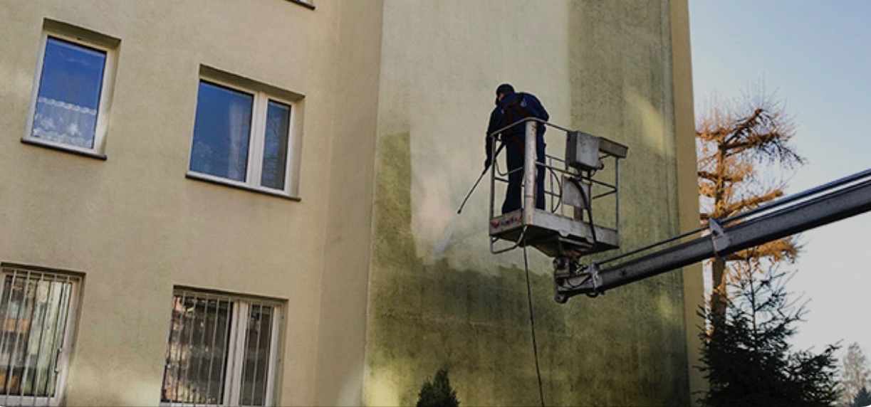 Pracownik na podnośniku koszowym czyści elewację budynku wielorodzinnego za pomocą myjki ciśnieniowej, widoczne różnice w kolorze elewacji przed i po czyszczeniu, zieleń porastająca budynek w tle.