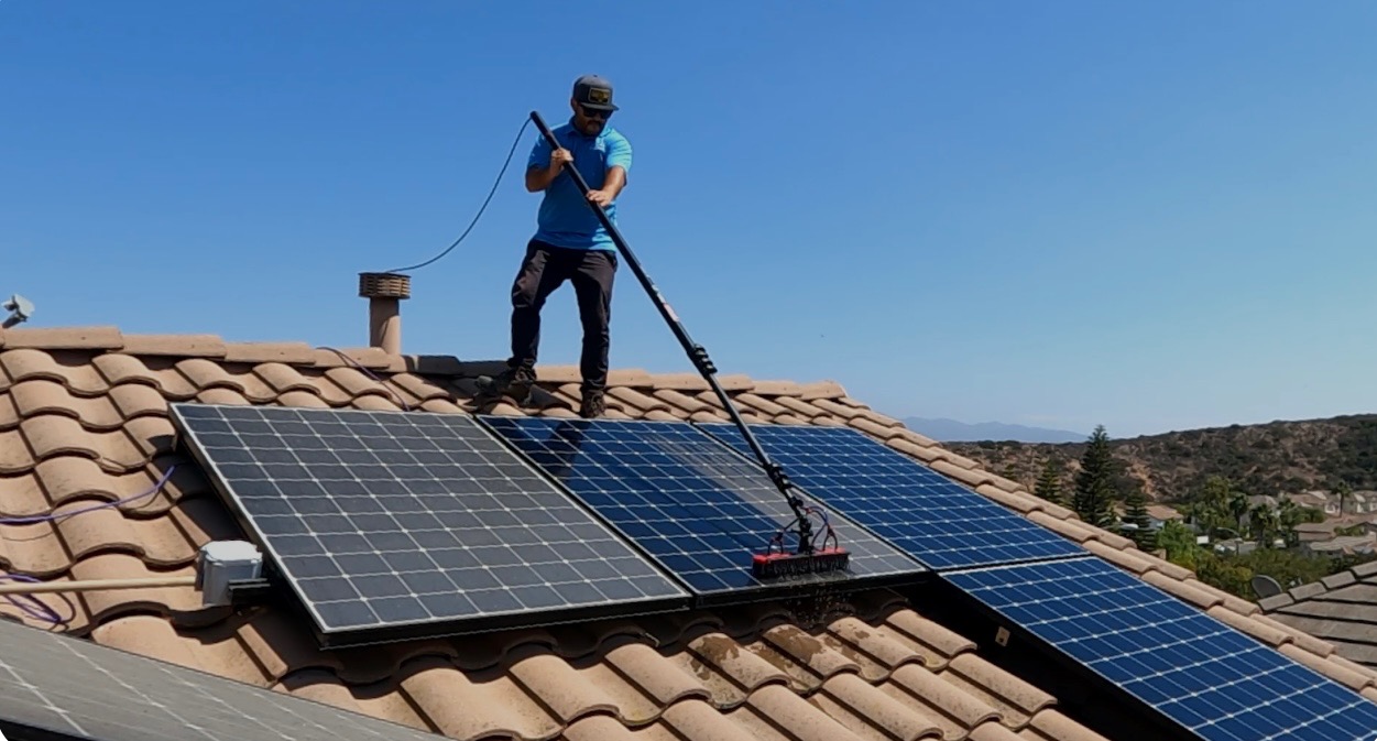 Osoba w okularach przeciwsłonecznych czyści panele słoneczne na dachu domu za pomocą szczotki na długim wysięgniku, na tle błękitnego nieba i zabudowań.