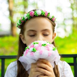 KUCIAFOTO - Dziewczynka z zamkniętymi oczami wącha bukiet z białych i różowych kwiatów, na głowie ma wianek z podobnych kwiatów.