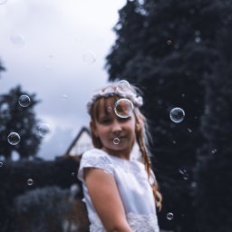 KUCIAFOTO - Dziewczynka w białej sukience i wianku na głowie, otoczona bańkami mydlanymi na tle domu i drzew