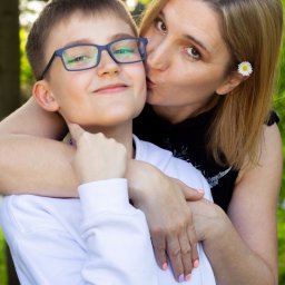 KUCIAFOTO - Portret matki całującej syna w policzek, on w okularach uśmiecha się, na włosach matki biały kwiatek, tło zielone.