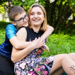 KUCIAFOTO - Uśmiechnięta kobieta w kwiecistej sukience siedzi na trawie, obejmowana przez chłopca w okularach i niebieskiej koszulce; w tle zieleń drzew i białe kwiaty.