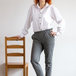 KUCIAFOTO - Kobieta z rudymi kręconymi włosami, w białej koszuli i szarych spodniach w paski, pozuje opierając się o drewniane krzesło na tle białej ściany.