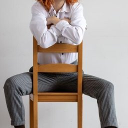 KUCIAFOTO - Kobieta z rudymi włosami, ubrana w białą koszulę i spodnie w paski, siedzi na drewnianym krześle, spoglądając w prawo, z rękami założonymi na oparciu krzesła.