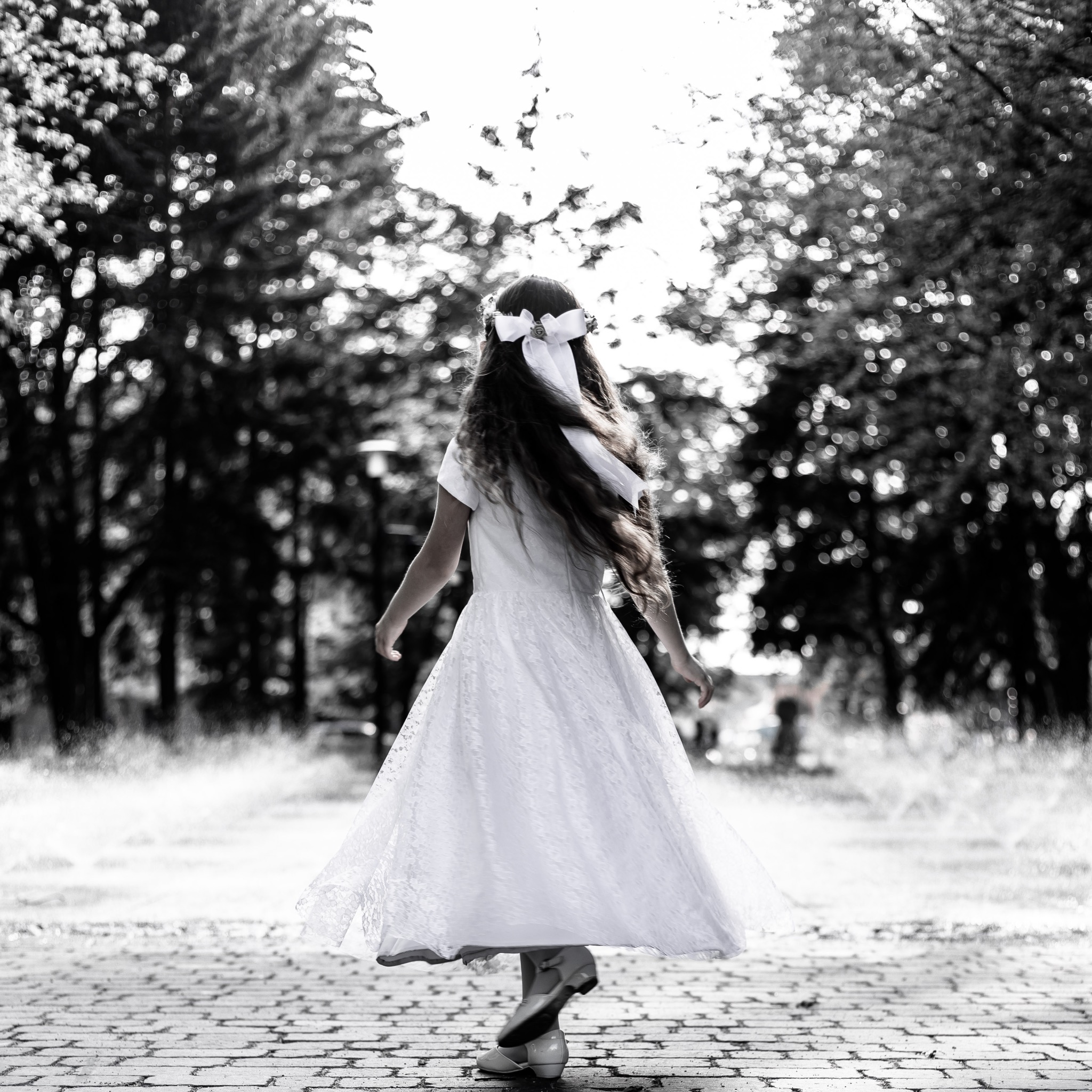 Dziewczynka w białej sukience i z kokardą we włosach, odwrócona tyłem, tańczy na brukowanej ścieżce w parku, w tle drzewa i rozproszone światło.