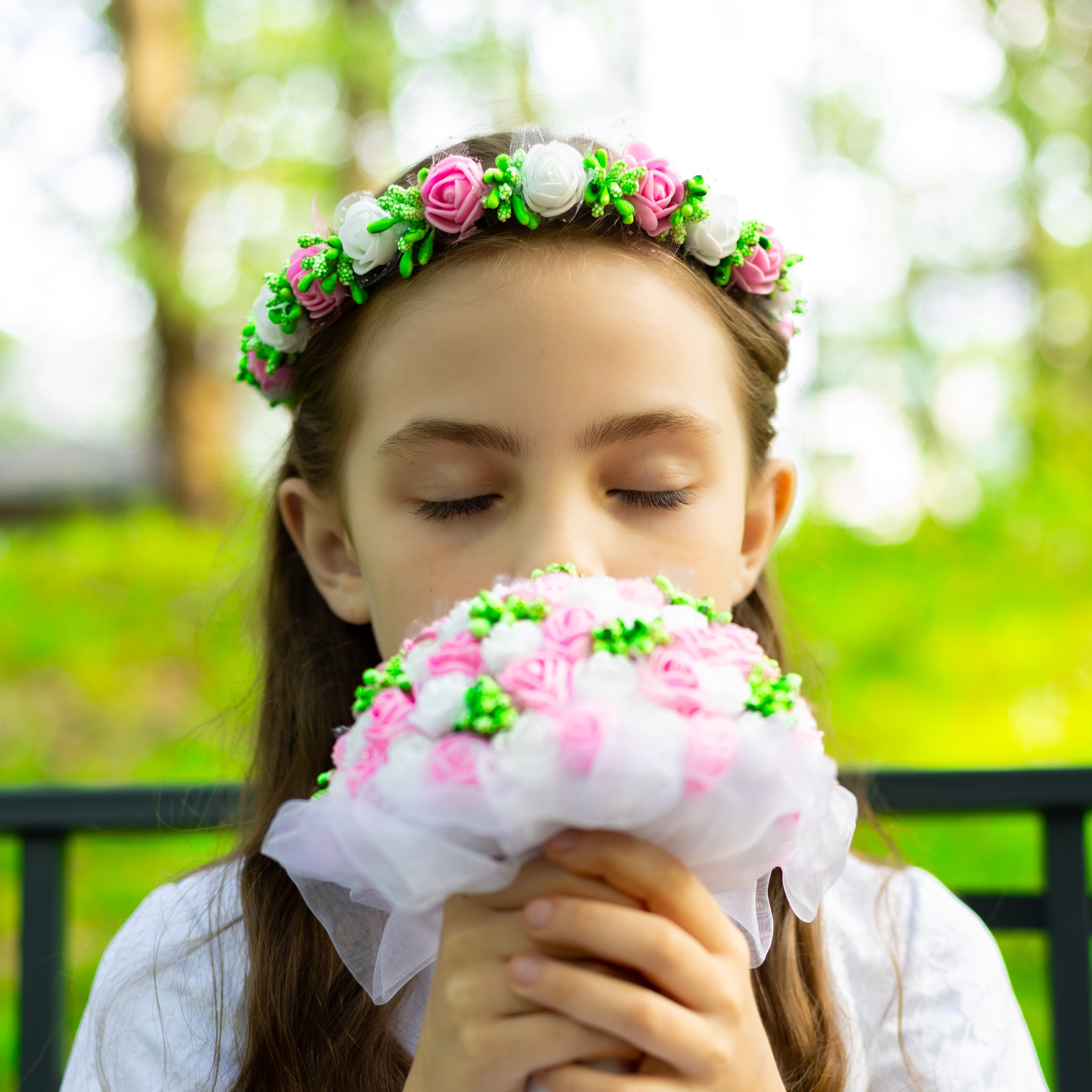 Dziewczynka z zamkniętymi oczami wącha bukiet z białych i różowych kwiatów, na głowie ma wianek z podobnych kwiatów.