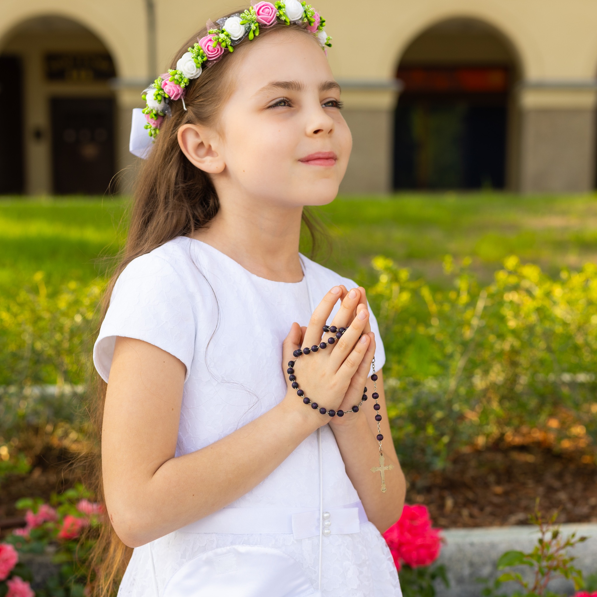 Dziewczynka w białej sukience i wianku na głowie, trzymająca różaniec, z zamyślonym wyrazem twarzy, na tle zieleni i budynku z arkadami.