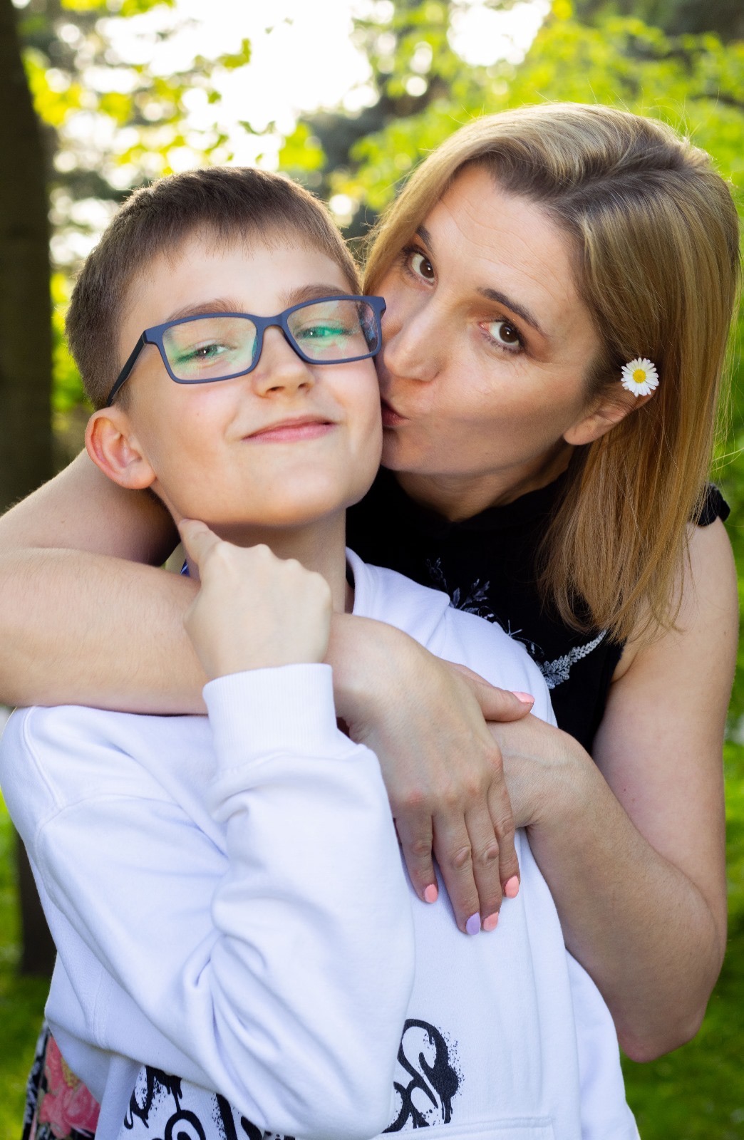 Portret matki całującej syna w policzek, on w okularach uśmiecha się, na włosach matki biały kwiatek, tło zielone.